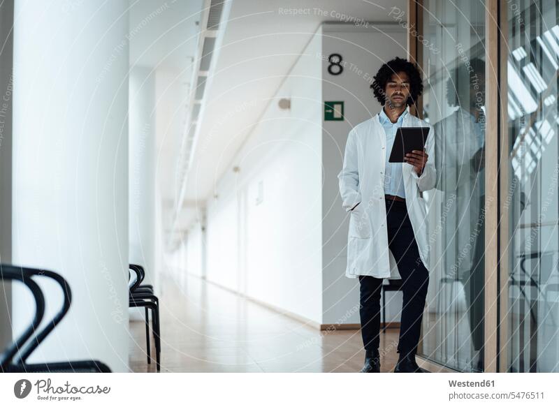 Confident young male doctor using digital tablet while leaning on glass wall at hospital corridor color image colour image Germany indoors indoor shot
