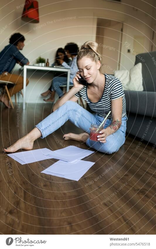 Young woman sitting on floor with cell phone and papers and friends in background floors Seated mobile phone mobiles mobile phones Cellphone cell phones