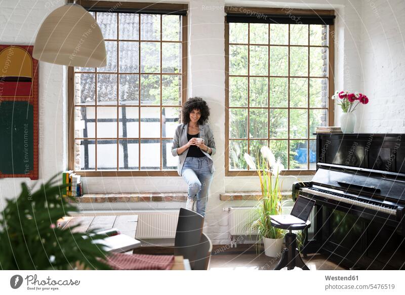 Woman standing at window of her loft apartment, drinking coffee windows Coffee lofts woman females women home at home piano pianos Drink beverages Drinks