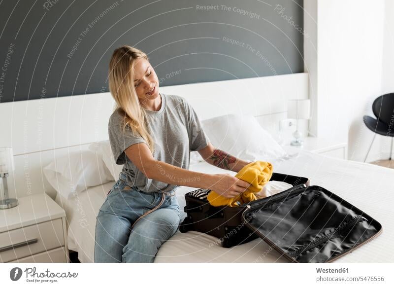 Smiling young woman sitting on bed with suitcase suitcases beds smiling smile Seated females women Adults grown-ups grownups adult people persons human being