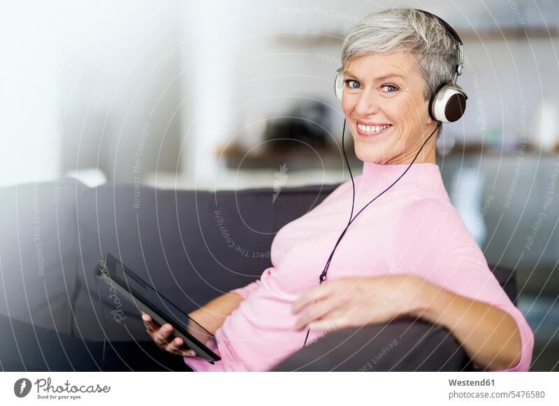 Portrait of happy mature woman sitting on couch with digital tablet listening music with headphones Seated portrait portraits females women happiness headset