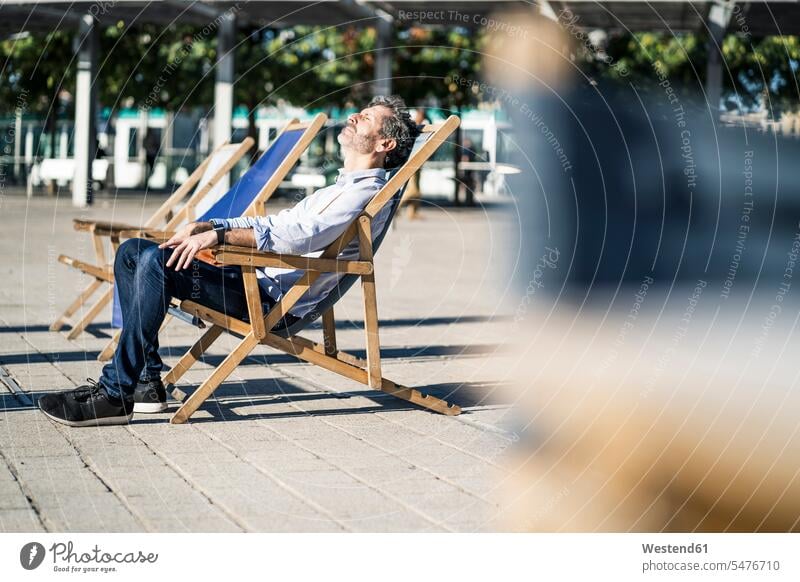 Mature man relaxing in deckchair on a square in the city town cities towns sitting Seated men males deck chair deckchairs deck chairs plaza places Public Square