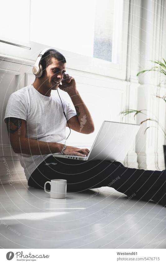 Smiling young man with headphones sitting on the floor using laptop smiling smile floors Seated men males Laptop Computers laptops notebook headset Adults