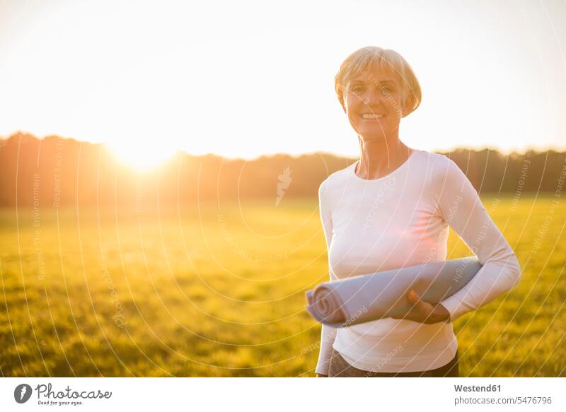 Portrait of smiling senior woman holding yoga mat on rural meadow at sunset country countryside portrait portraits sunsets sundown females women senior women