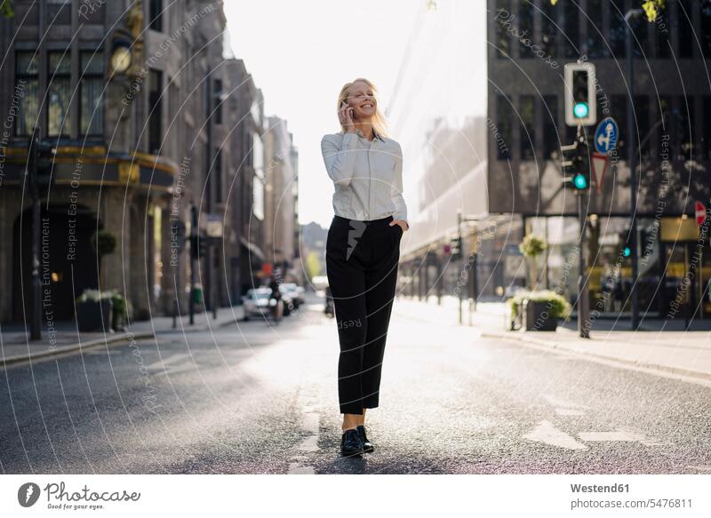 Smiling businesswoman talking through mobile phone while looking away and walking on street in city color image colour image Germany outdoors location shots