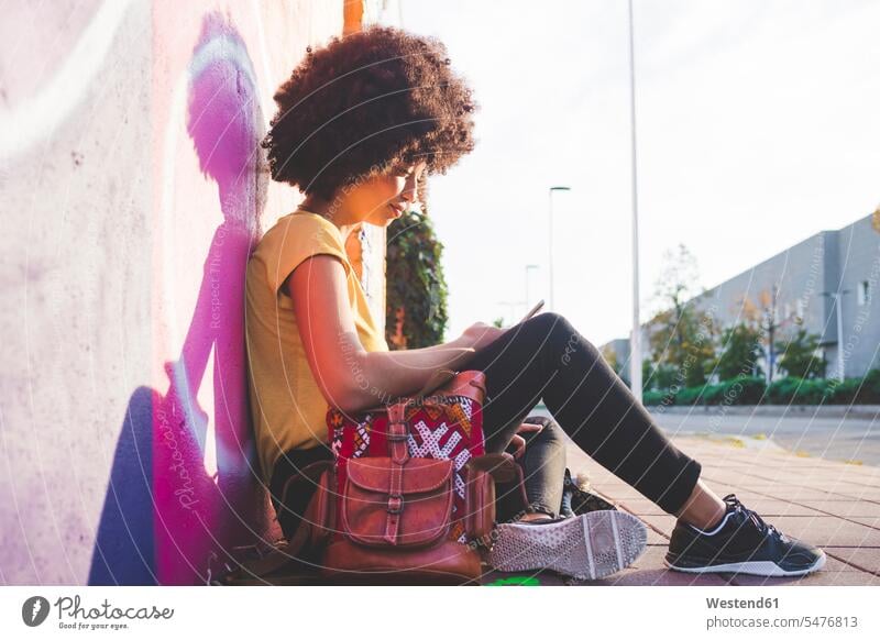 Young woman with afro hairdo sitting at graffiti wall and using smartphone human human being human beings humans person persons curl curled curls curly hair