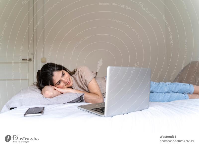 Smiling young woman lying on bed with smartphone and laptop human human being human beings humans person persons caucasian appearance caucasian ethnicity