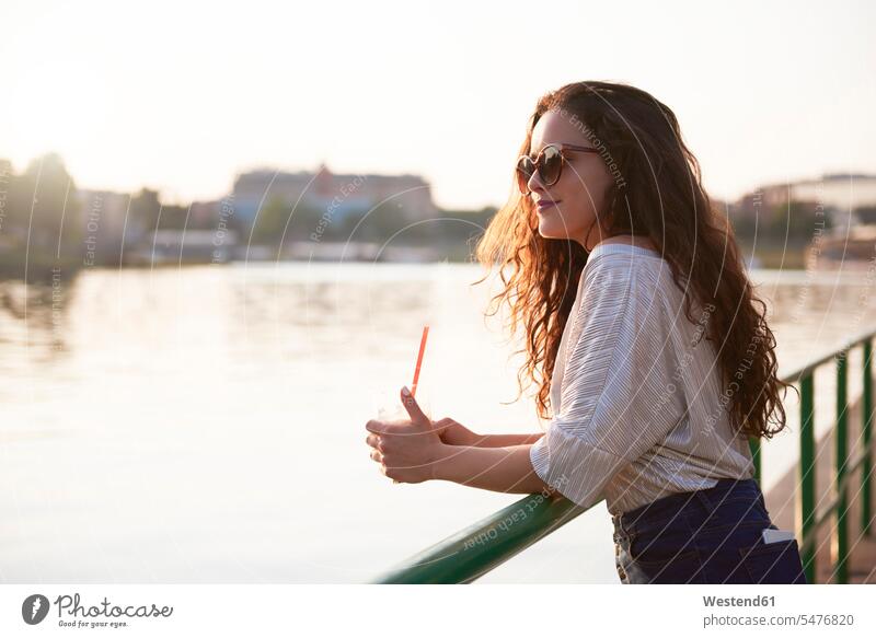Smiling young woman with a takeaway drink enjoying the sunset at the riverside Drink beverages Drinks Beverage sunsets sundown indulgence enjoyment savoring