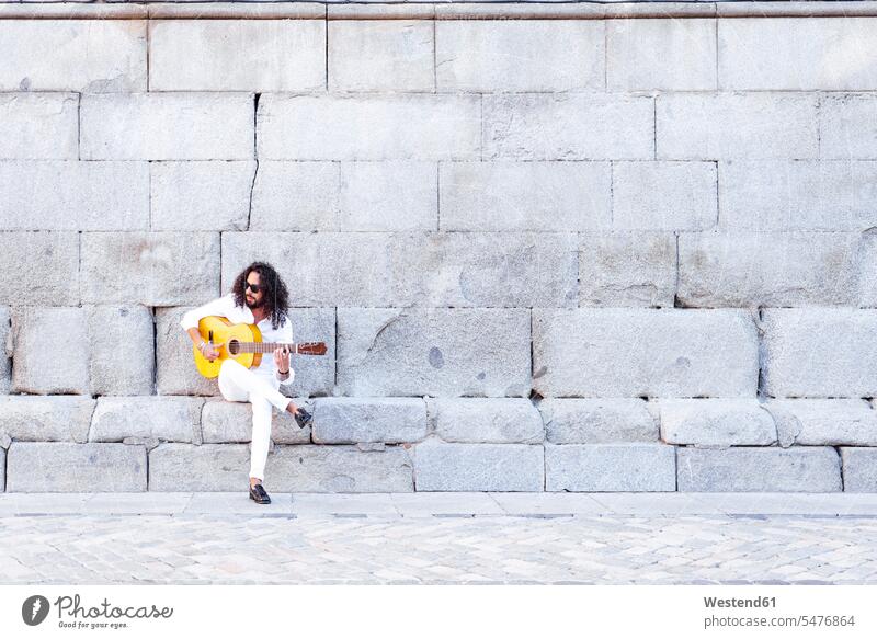 Guitarist playing guitar while sitting against concrete wall color image colour image outdoors location shots outdoor shot outdoor shots day daylight shot