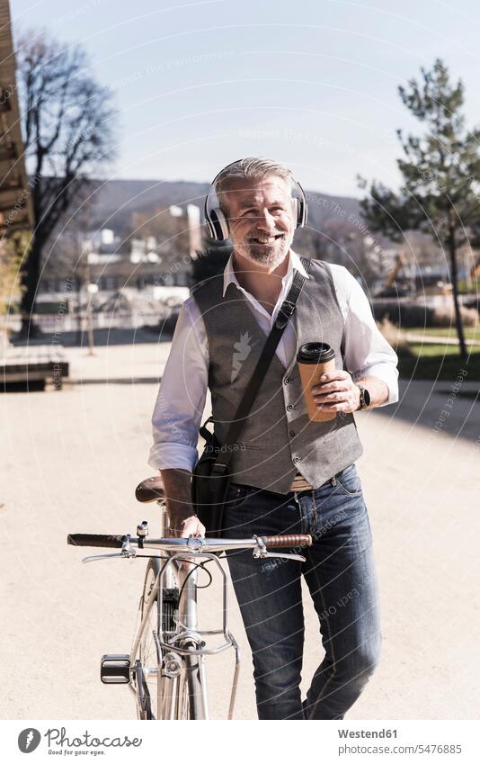 Smiling mature businessman with bicycle, takeaway coffee and headphones on the go in the city smiling smile Businessman Business man Businessmen Business men
