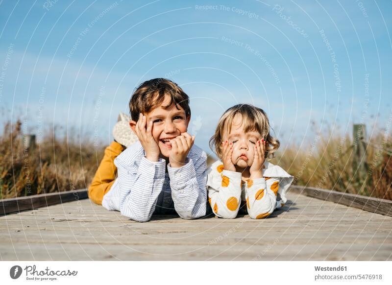 Portrait of boy and his little sister lying side by side on boardwalk pulling funny faces Harmony Harmonious paralell Juxtaposed in paralell sisters brother