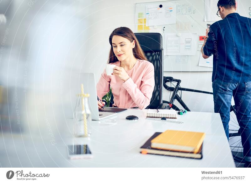 Woman working at desk in office with colleague in background At Work colleagues offices office room office rooms desks workplace work place place of work Table