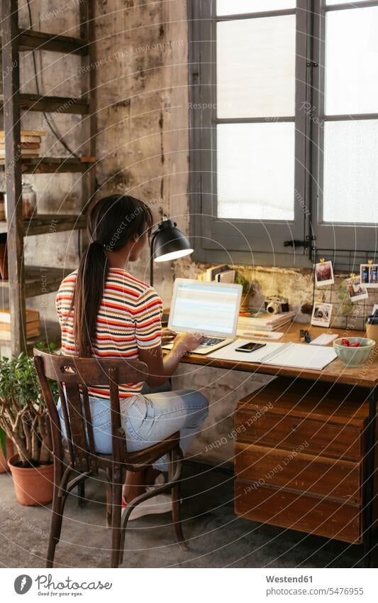 Back view of young woman sitting at desk in a loft working on laptop Laptop Computers laptops notebook Seated lofts females women desks At Work computer