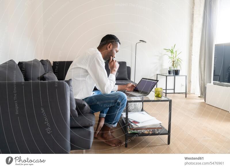 Man sitting on couch in modern apartment using laptop books couches settee settees sofa sofas computers Laptop Computer Laptop Computers laptops notebook Seated
