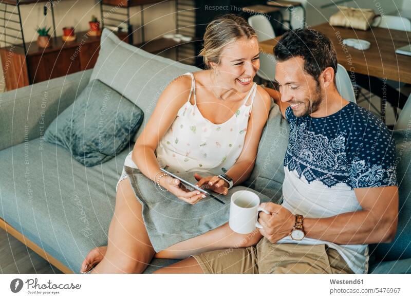 Happy relaxed couple sitting on couch in living room sharing a tablet cushions couches settee settees sofa sofas hold smile Seated speak speaking talk drink