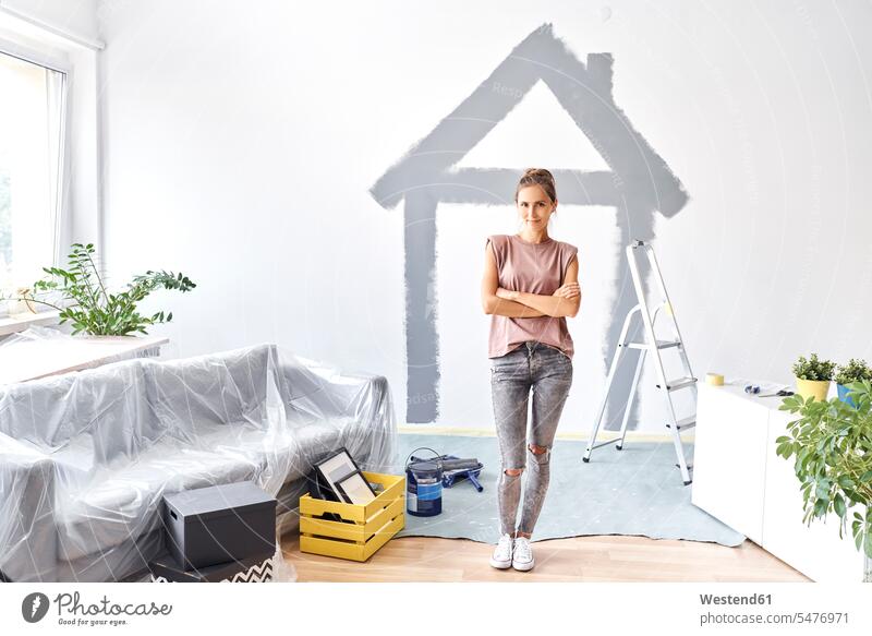 Young woman with arms crossed standing against house painted on wall at home color image colour image indoors indoor shot indoor shots interior interior view