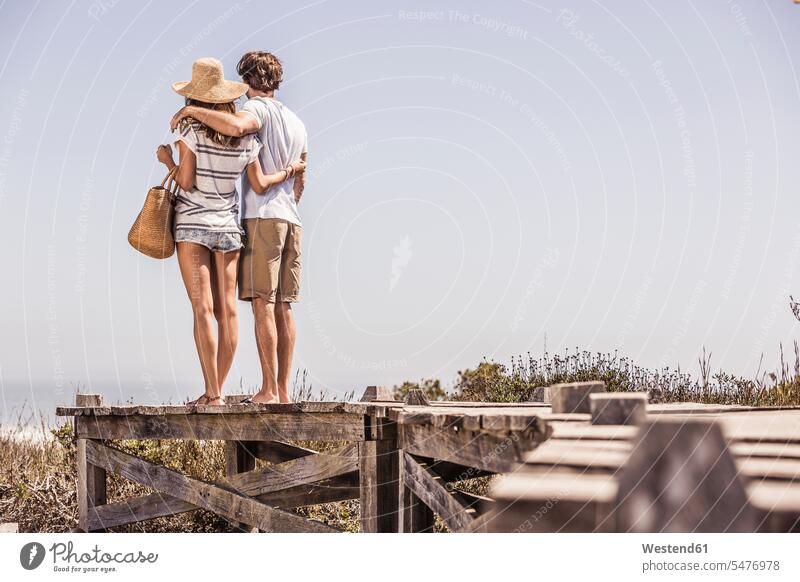Young couple standing on a boardwalk at the coast looking at view touristic tourists bags embrace Embracement hug hugging seasons summer time summertime summery