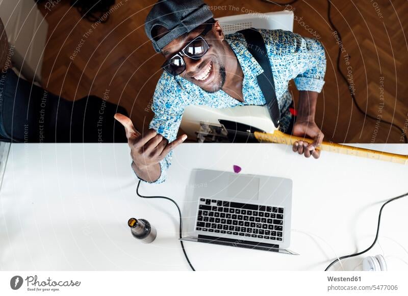 Portrait of smiling young man showing shaka sign while sitting with guitar and laptop at desk color image colour image indoors indoor shot indoor shots interior