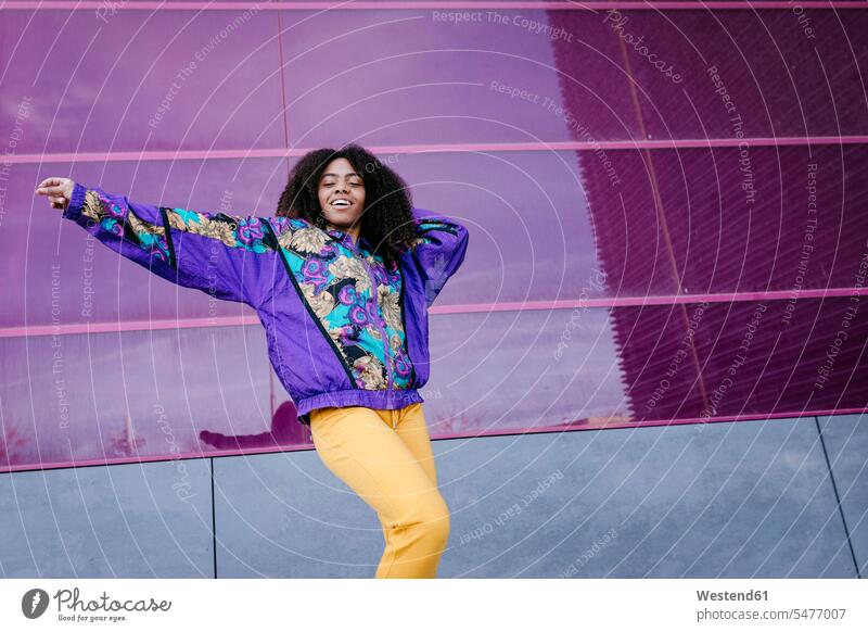 Young woman with urban look dancing, pink glass wall in the background coat coats jackets smile Ardor Ardour enthusiasm enthusiastic excited delight enjoyment