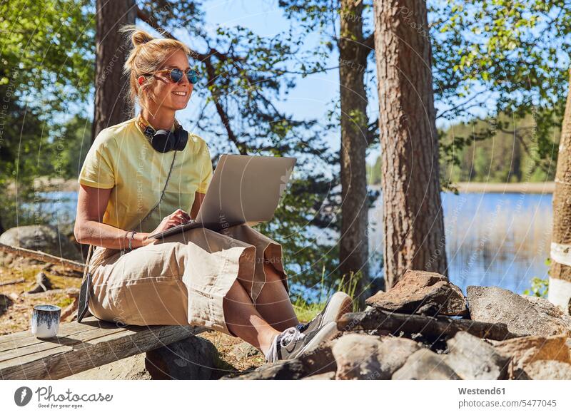 Mid adult woman wearing sunglasses using laptop while sitting in Tiveden National Park, Sweden color image colour image Scandinavia Scandinavian Peninsula