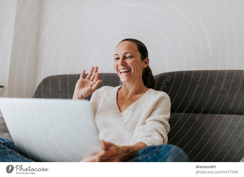 Happy woman waving hand video call on laptop at home color image colour image Connection connected connectivity Connections domestic life domestic scene