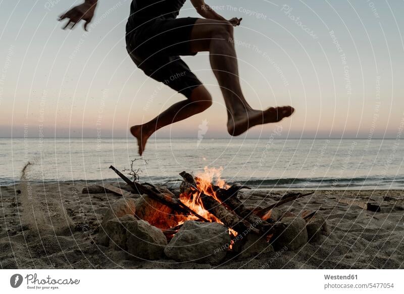Man jumping over campfire at beach during sunset color image colour image outdoors location shots outdoor shot outdoor shots sunsets sundown atmosphere Idyllic