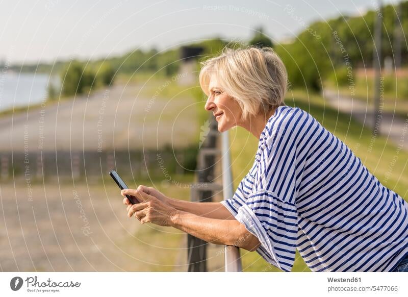 Smiling senior woman with cell phone leaning on railing at riverbank mobile phone mobiles mobile phones Cellphone cell phones riverside females women smiling