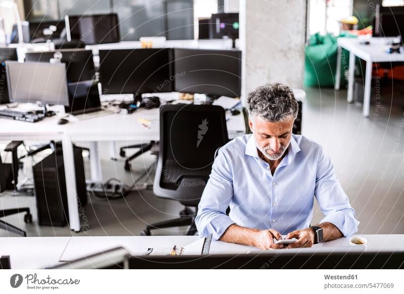 Mature businessman sitting at desk in office using smartphone desks Seated Smartphone iPhone Smartphones offices office room office rooms Businessman