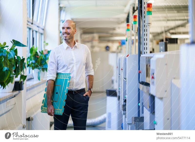Smiling male engineer looking away while holding large circuit board by machinery at factory color image colour image indoors indoor shot indoor shots interior