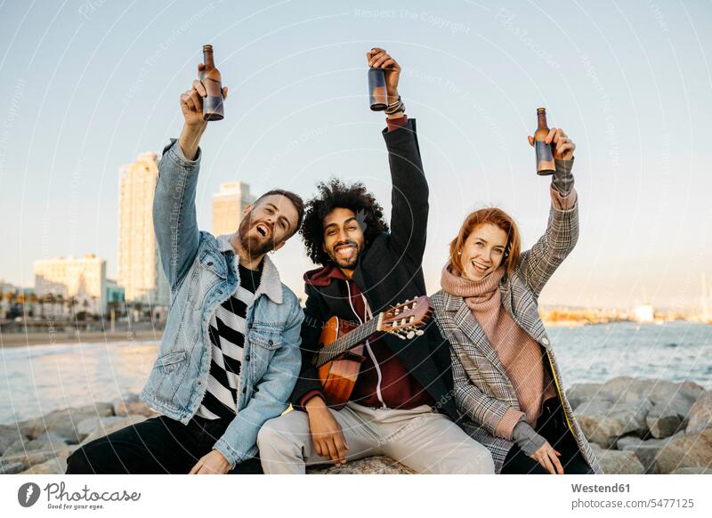 Portrait of three happy friends with guitar raising beer bottles at the coast sunset coastline shoreline happiness sunsets sundown portrait portraits sitting