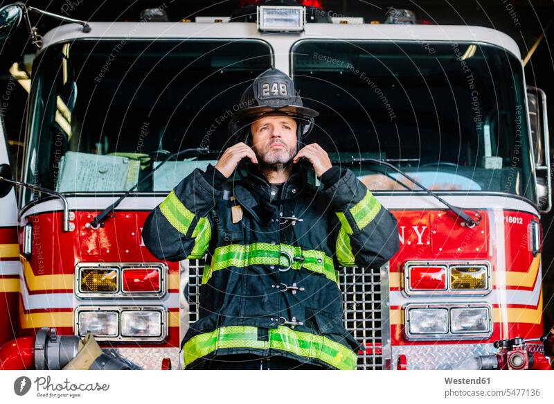 USA, New York, Portrait of firefighter putting on helmet in front of fire engine United States of America portrait portraits testimonial portrait front view