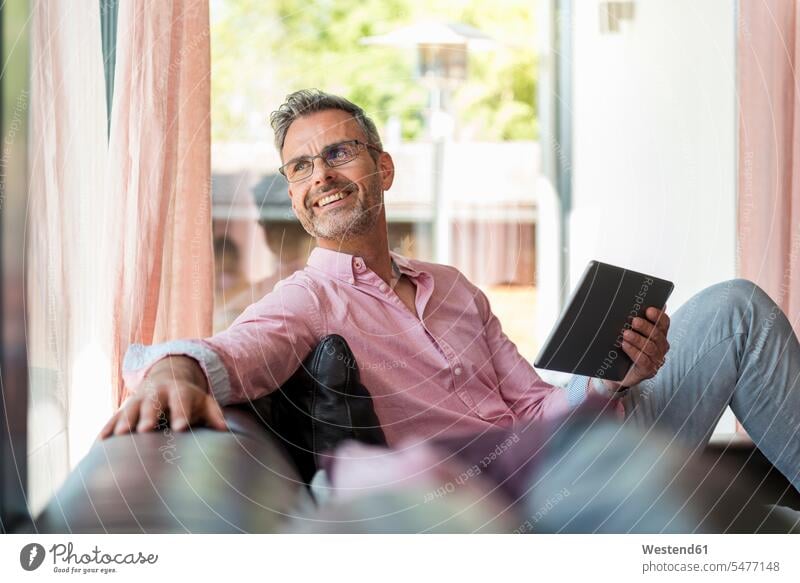 Smiling mature man sitting on couch at home holding a tablet Seated smiling smile settee sofa sofas couches settees men males digitizer Tablet Computer