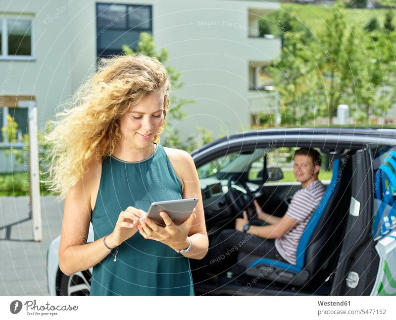 Smiling young woman using tablet with man sitting in electric car in background automobile Auto cars motorcars Automobiles smiling smile couple twosomes