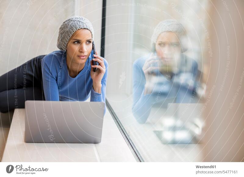 Portrait of young woman on the phone with laptop on window sill looking out of window watching Laptop Computers laptops notebook females women portrait