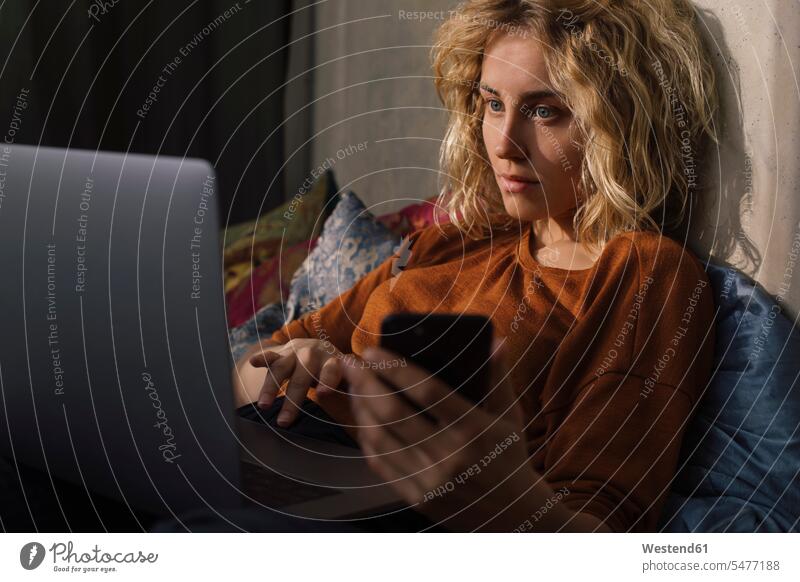 Portrait of blond young woman lying on bed using cell phone and laptop human human being human beings humans person persons caucasian appearance