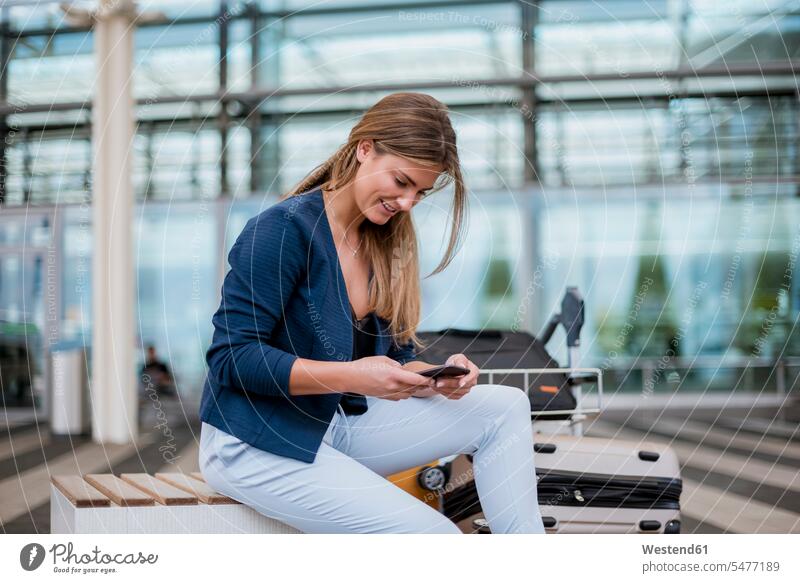 Smiling young businesswoman sitting outdoors with cell phone and suitcase suitcases mobile phone mobiles mobile phones Cellphone cell phones businesswomen