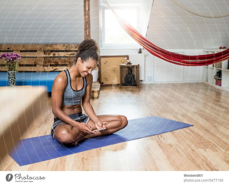 Smiling young woman sitting on yoga mat looking at cell phone females women Yoga Mat eyeing mobile phone mobiles mobile phones Cellphone cell phones smiling