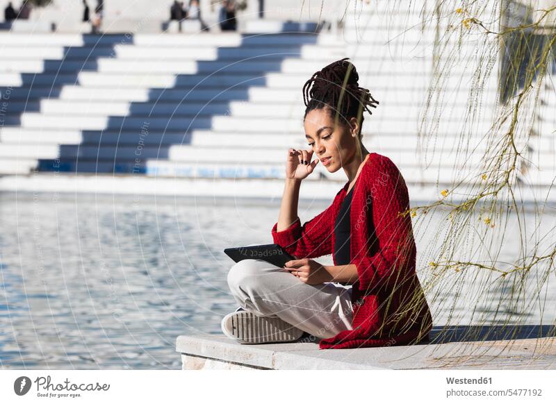 Young woman sitting cross-legged using digital tablet while at edge of promenade over river on sunny day color image colour image Spain outdoors location shots