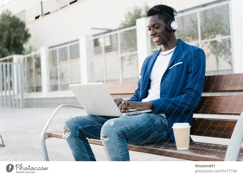 Smiling young businessman using laptop while listening music on bench at footpath color image colour image outdoors location shots outdoor shot outdoor shots