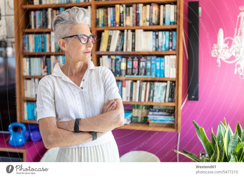 Senior female with arms crossed standing against bookshelf at home color image colour image day daylight shot daylight shots day shots daytime casual clothing