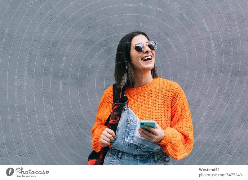 Smiling woman with sunglasses and purse using mobile phone while standing against gray wall color image colour image outdoors location shots outdoor shot