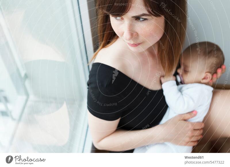 Young mother with her baby girl looking out of the window at home windows comforter Pacifiers soother hold Seated sit Secure closeness propinquity contemplative