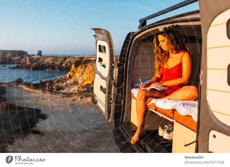 Woman writing in book while sitting in camper van against at beach color image colour image outdoors location shots outdoor shot outdoor shots sunset sunsets