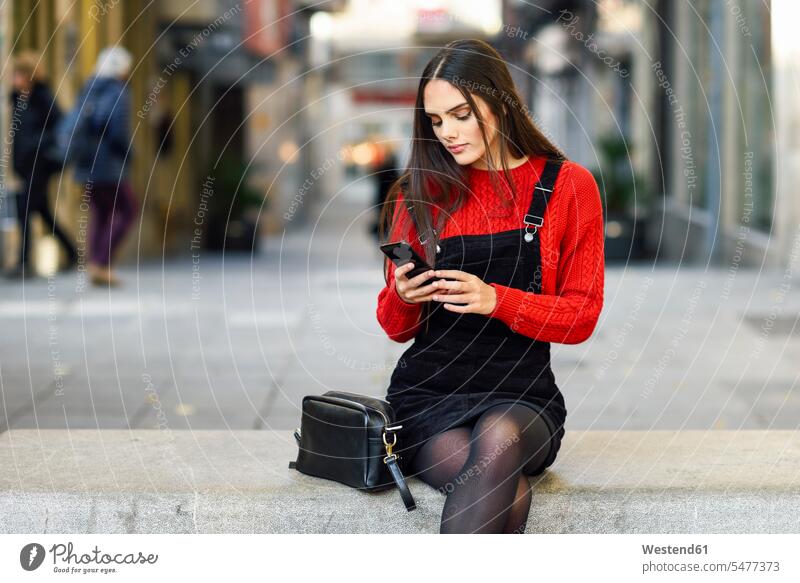 Portrait of fashionable young woman sitting on bench at pedestrian area looking at cell phone Seated females women Smartphone iPhone Smartphones eyeing portrait