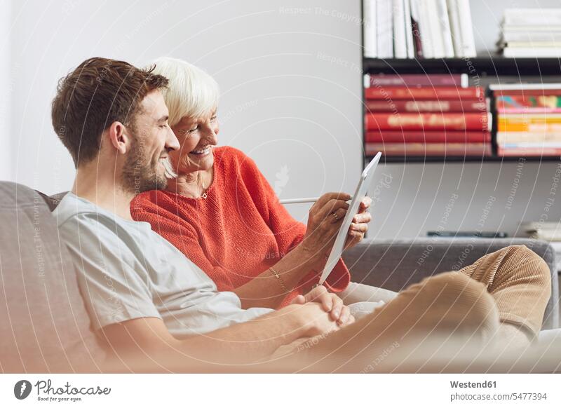Grandson sitting on couch, using digital tablet with his grandmother human human being human beings humans person persons adult grown-up grown-ups grownup