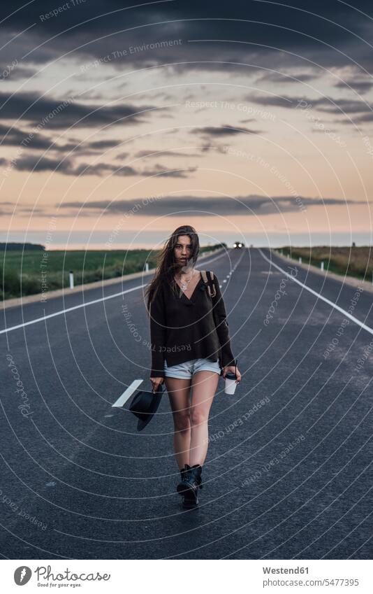 Portrait of hitchhiking young woman with backpack and beverage walking on lane at evening twilight portrait portraits Lane Roadway females women hitchhike