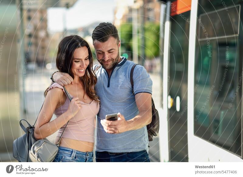 Couple looking at smartphone while waiting for tram at the station mobile phone mobiles mobile phones Cellphone cell phone cell phones eyeing tramway tramways