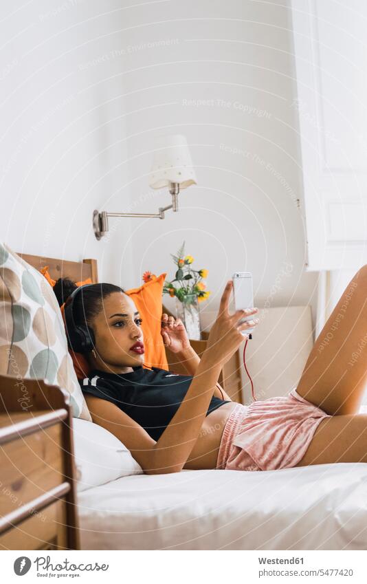 Beautiful young woman lying on bed at home wearing headphones looking at cell phone eyeing beds headset mobile phone mobiles mobile phones Cellphone cell phones