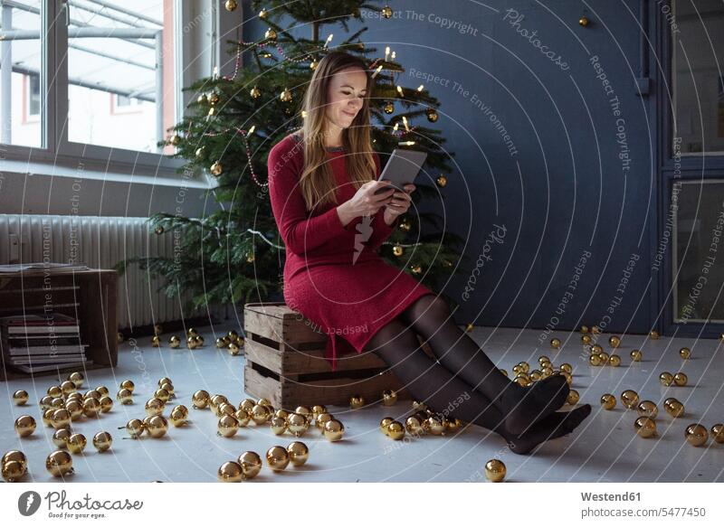Smiling woman sitting on wooden box in front of decorated Christmas tree using tablet females women Seated digitizer Tablet Computer Tablet PC Tablet Computers