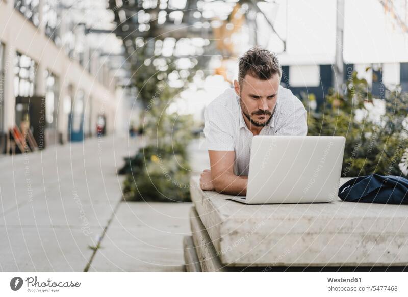Young man with laptop and looking sideways, lying on a bench in the city Occupation Work job jobs profession professional occupation business life